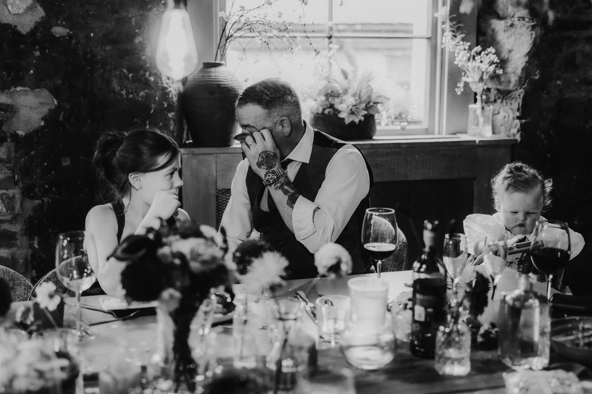 Man wiping his eyes at a wedding table with two children beside him, surrounded by glasses and flowers.