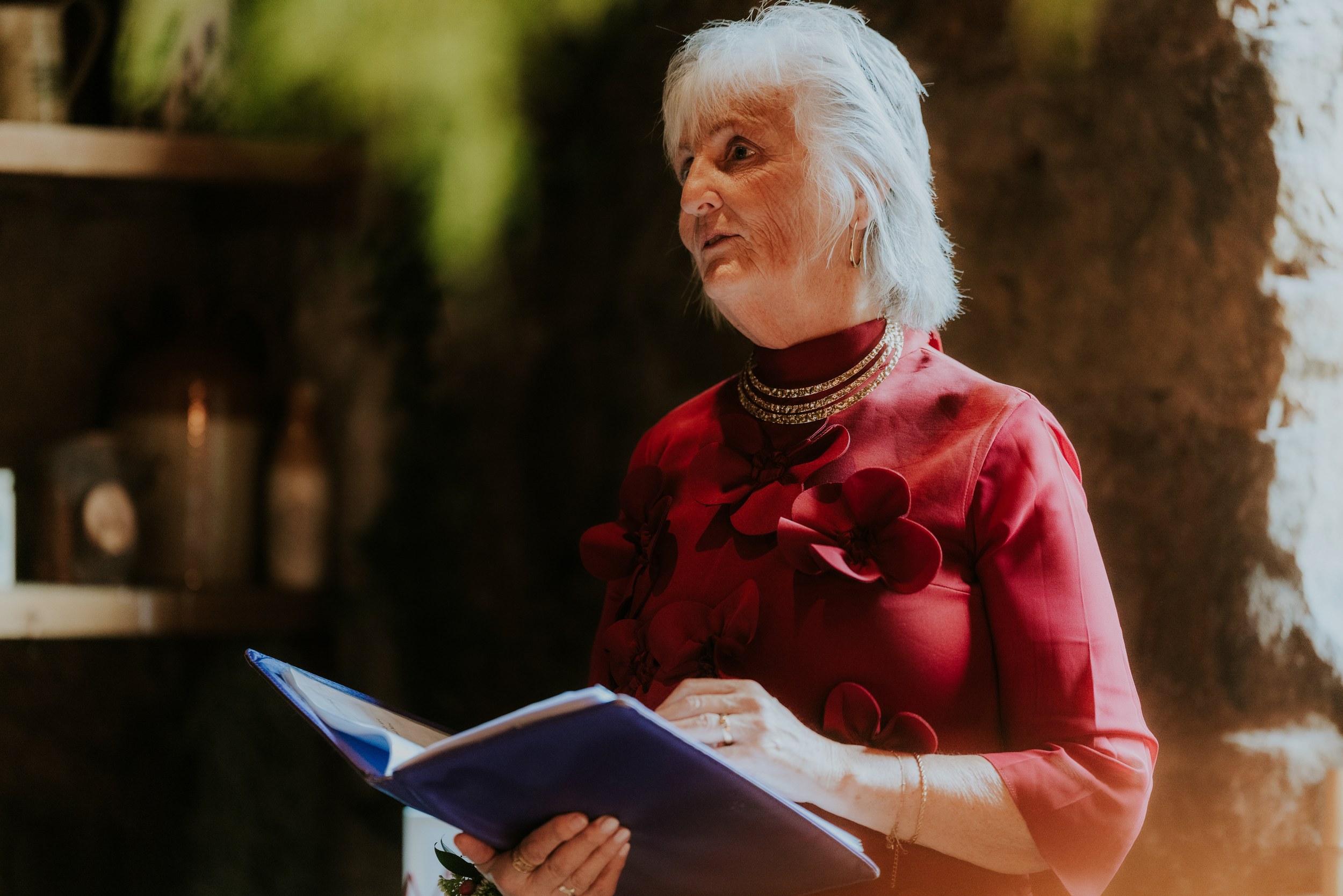 Older woman in a red dress reading from a blue folder and speaking indoors at the wedding.