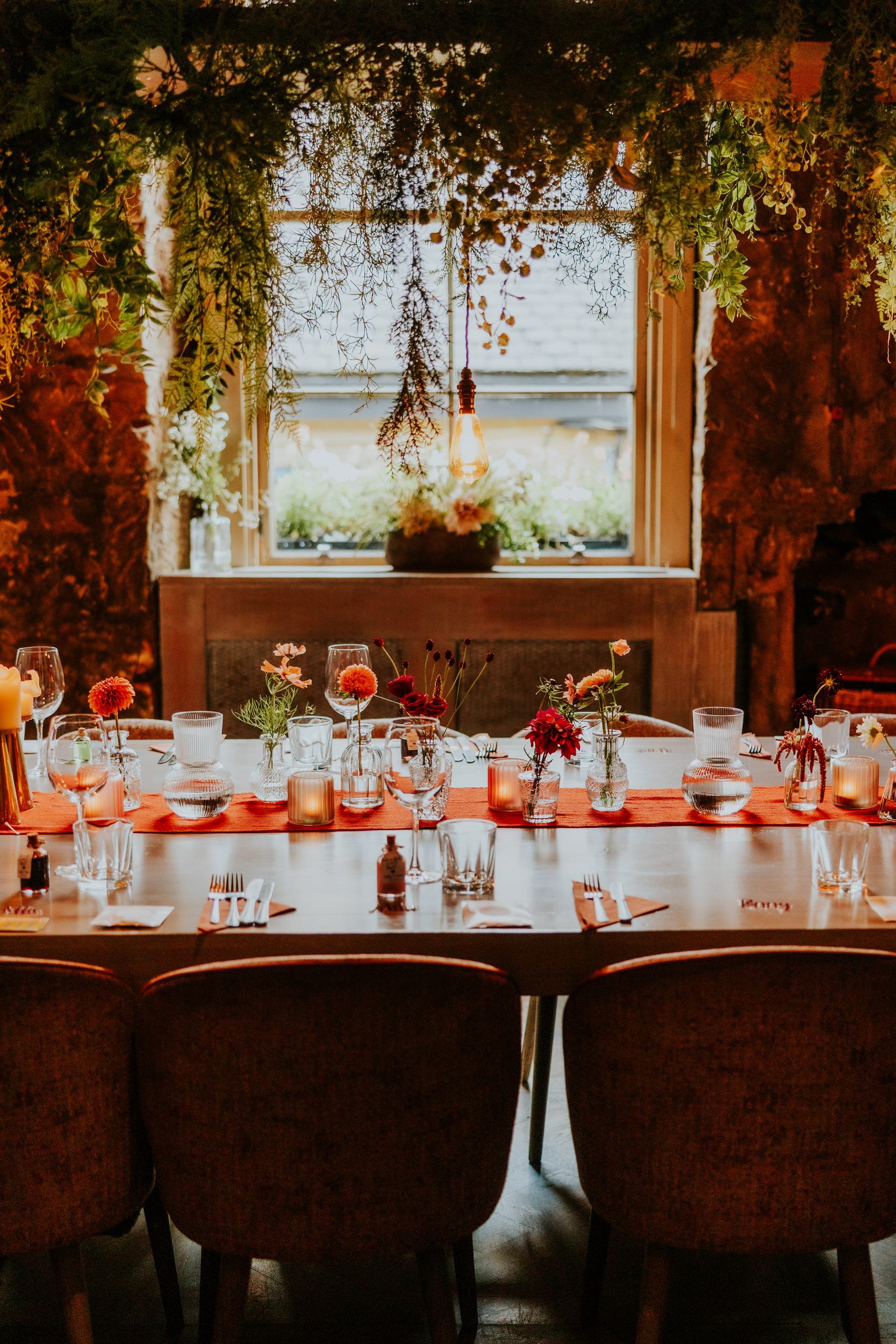 Wedding reception table with glassware, small floral vases and candles under hanging greenery and an exposed bulb by a window.