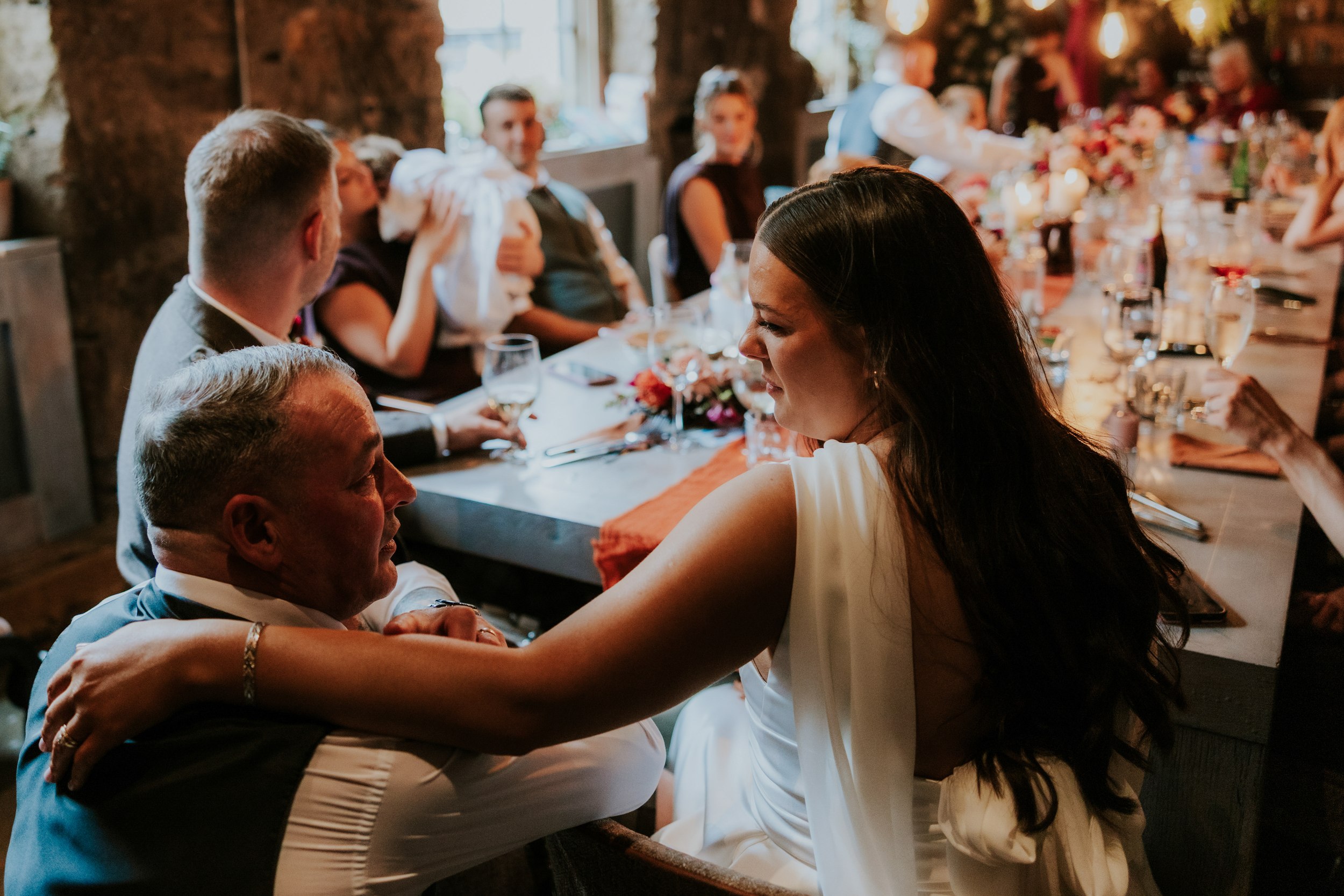 Woman in a white dress leans over to embrace a seated guest at a long, candlelit wedding reception table.