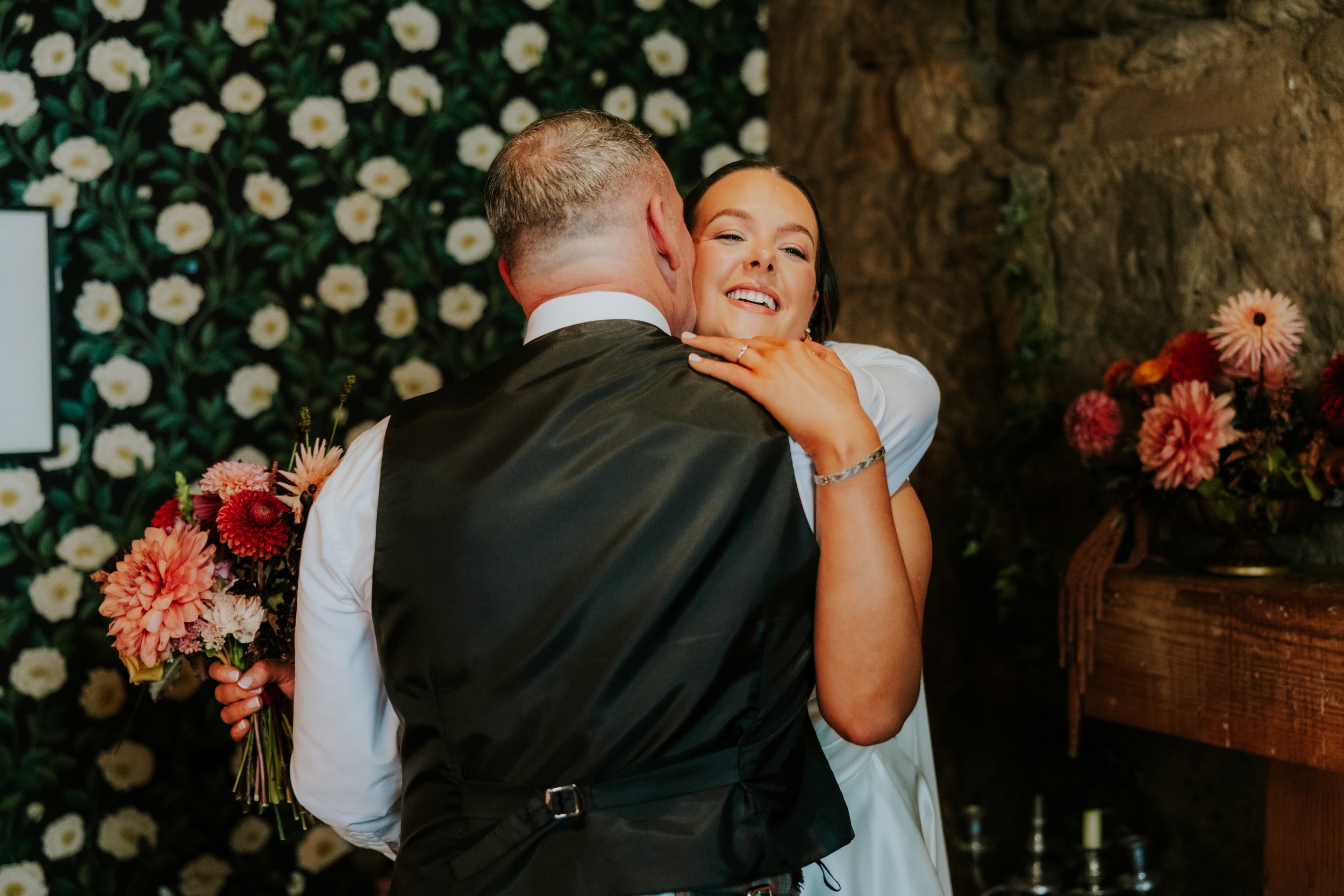 Woman in a white dress smiling as she hugs a man in a dark waistcoat with bouquet and floral wallpaper behind them.