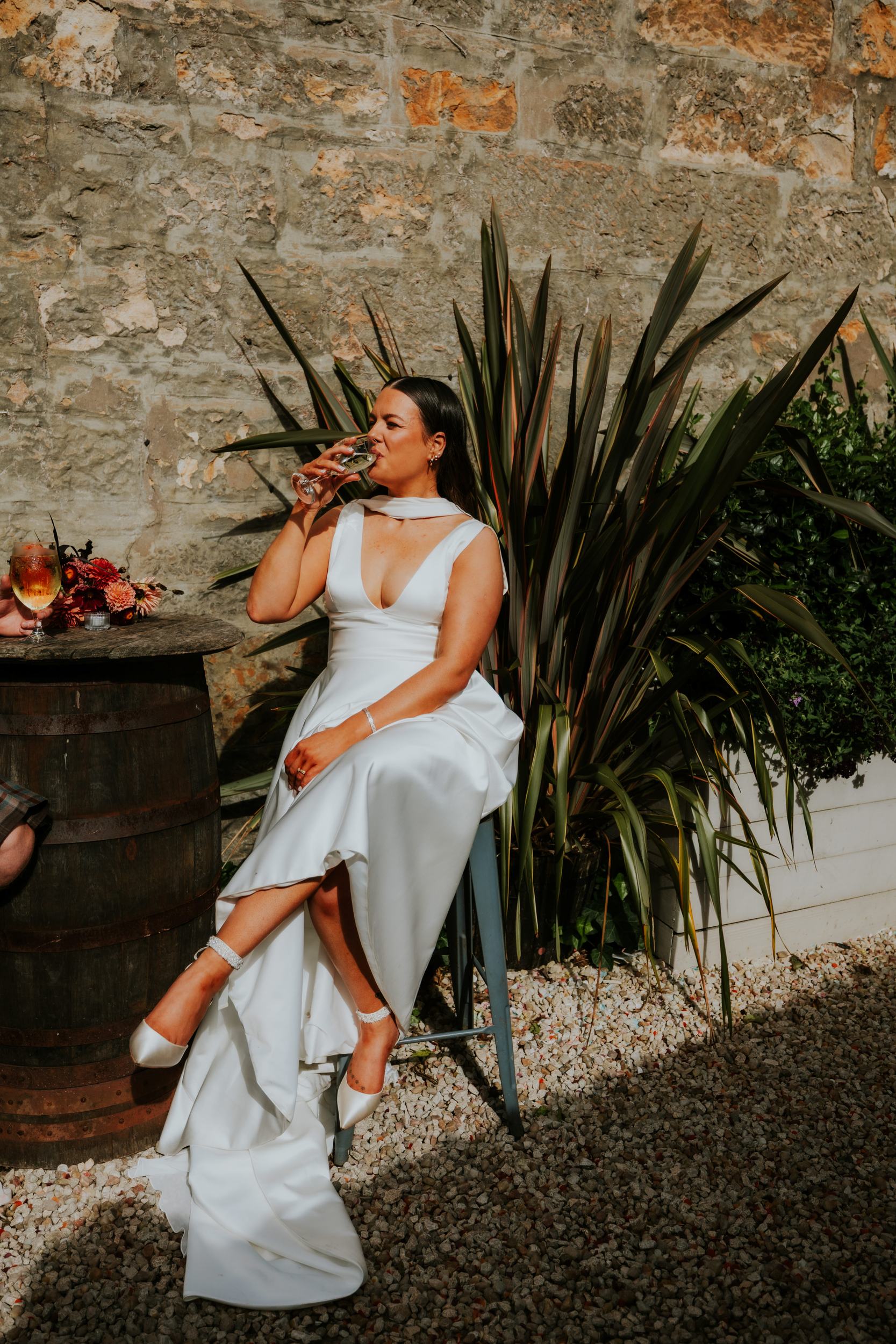 Woman in white wedding dress seated outdoors against a stone wall, sipping a glass of wine.