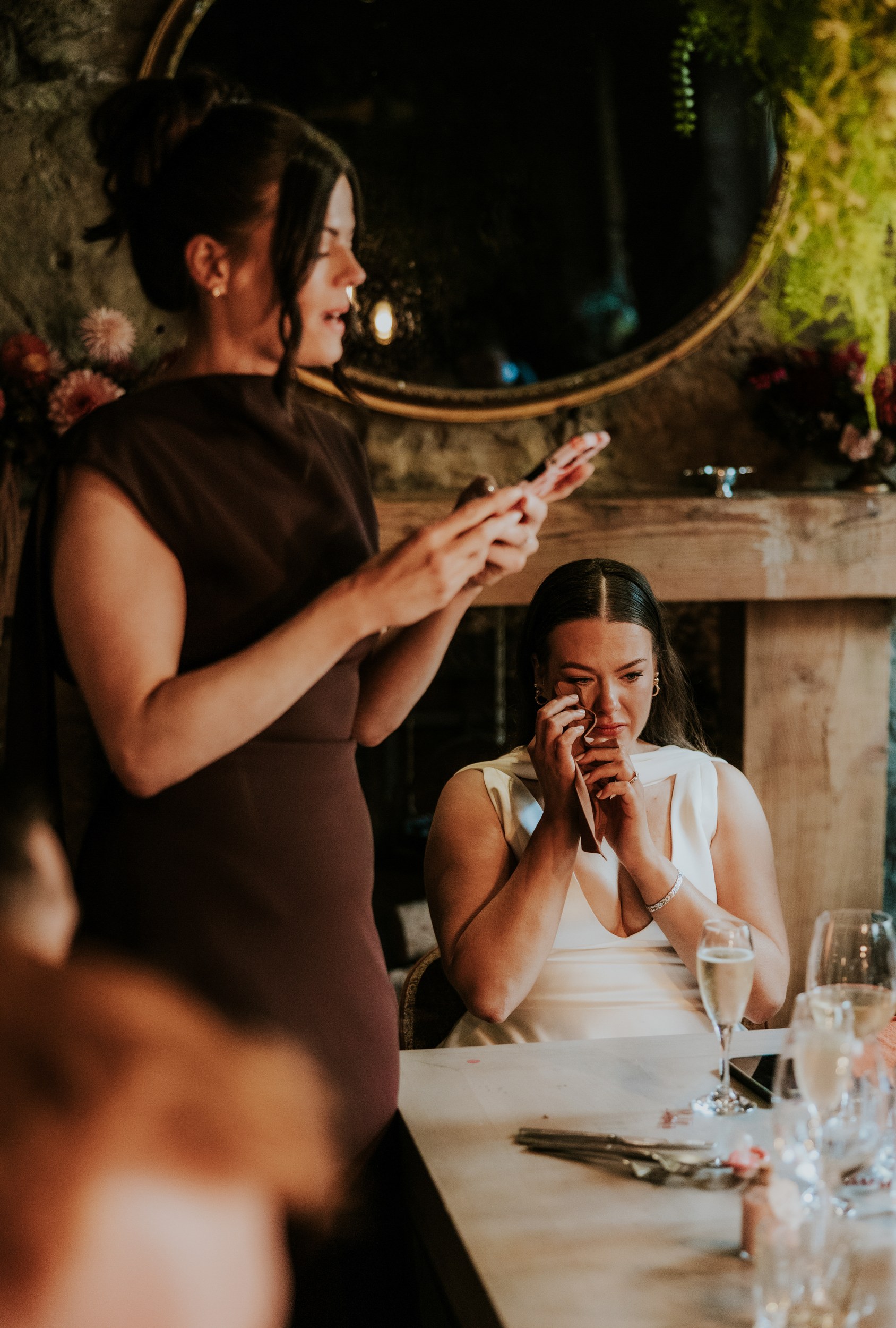 Woman reads from her phone during a wedding speech as a seated woman in white wipes away tears beside a table with champagne.